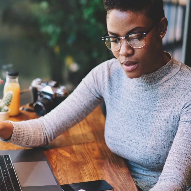 Une personne portant des lunettes travaille à une table en bois, regarde un écran d'ordinateur portable et écrit dans un cahier, avec un smartphone à proximité, suggérant un environnement de travail productif