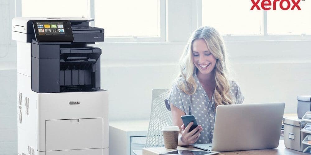 Une personne regarde joyeusement son smartphone, assise &agrave; un bureau avec un ordinateur portable. Une imprimante multifonction Xerox VersaLink B605 est visible &agrave; c&ocirc;t&eacute; du bureau, dans un espace de travail lumineux.