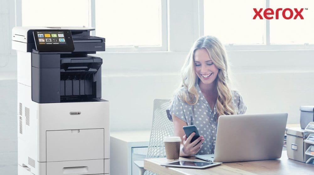 Une personne regarde joyeusement son smartphone, assise &agrave; un bureau avec un ordinateur portable. Une imprimante multifonction Xerox VersaLink B605 est visible &agrave; c&ocirc;t&eacute; du bureau, dans un espace de travail lumineux.