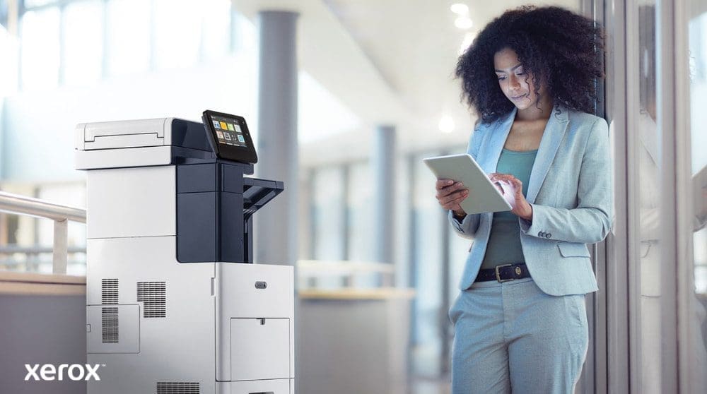 Une personne se tient &agrave; c&ocirc;t&eacute; d'une imprimante multifonction Xerox VersaLink B605, regardant et interagissant avec une tablette, illustrant comment ConnectKey permet une interaction mobile avec l'imprimante.