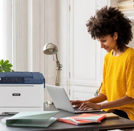 Une personne aux cheveux boucl&eacute;s sourit tout en tapant sur un ordinateur portable &agrave; un bureau, avec une imprimante compacte Xerox C310 positionn&eacute;e &agrave; c&ocirc;t&eacute; de l'ordinateur portable, pr&eacute;sentant une configuration de bureau &agrave; domicile moderne pour la productivit&eacute;.