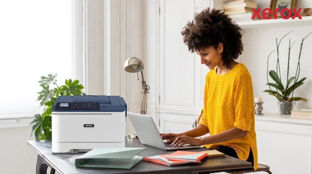 Une personne aux cheveux bouclés sourit tout en tapant sur un ordinateur portable à un bureau, avec une imprimante compacte Xerox C310 positionnée à côté de l'ordinateur portable, présentant une configuration de bureau à domicile moderne pour la productivité.