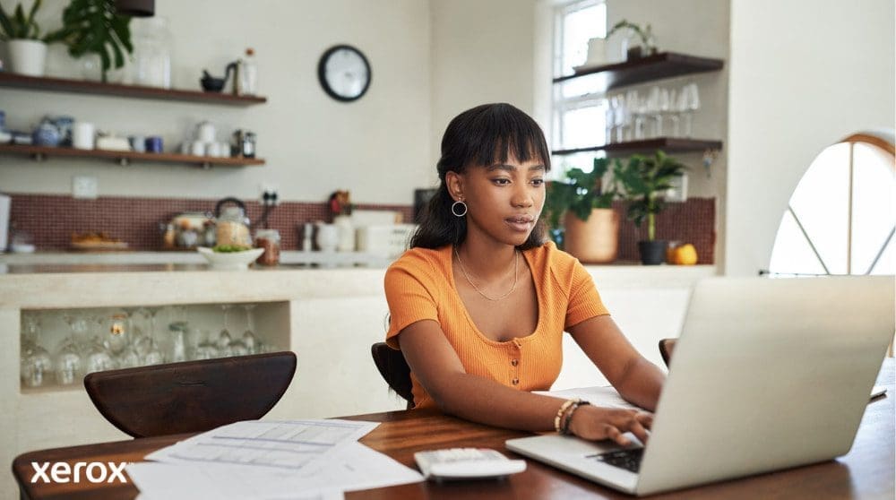 Une personne est assise &agrave; une table de cuisine, concentr&eacute;e sur son travail sur un ordinateur portable, avec des papiers et une calculatrice &agrave; proximit&eacute;, illustrant la flexibilit&eacute; du travail &agrave; domicile avec une gestion efficace des documents.