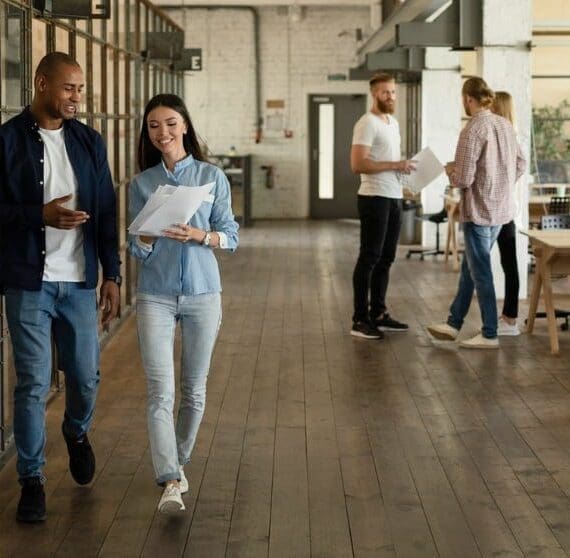Deux coll&egrave;gues marchent dans un couloir d'un bureau moderne et ouvert, tenant des documents et souriant, avec d'autres coll&egrave;gues visibles en arri&egrave;re-plan, repr&eacute;sentant un lieu de travail dynamique et collaboratif.