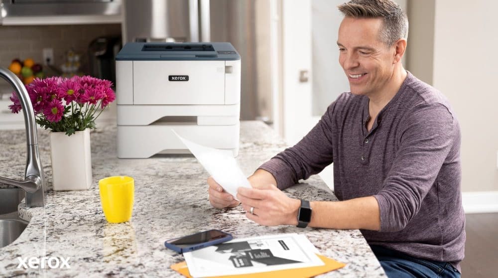 Une personne sourit tout en examinant un document sur un comptoir de cuisine, avec une imprimante Xerox compacte et une tasse jaune vif &agrave; proximit&eacute;, sugg&eacute;rant une impression et une gestion de documents transparentes dans un environnement domestique ou de petit bureau.