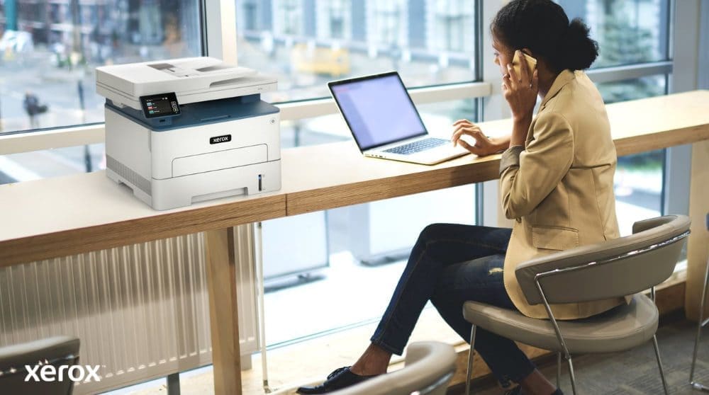 Une personne est assise &agrave; une table haute pr&egrave;s d'une grande fen&ecirc;tre, parlant au t&eacute;l&eacute;phone tout en travaillant sur un ordinateur portable. Une imprimante multifonction Xerox B235DNI est judicieusement plac&eacute;e sur la table &agrave; c&ocirc;t&eacute; d'elle, sugg&eacute;rant une utilisation simple dans un espace de travail moderne.
