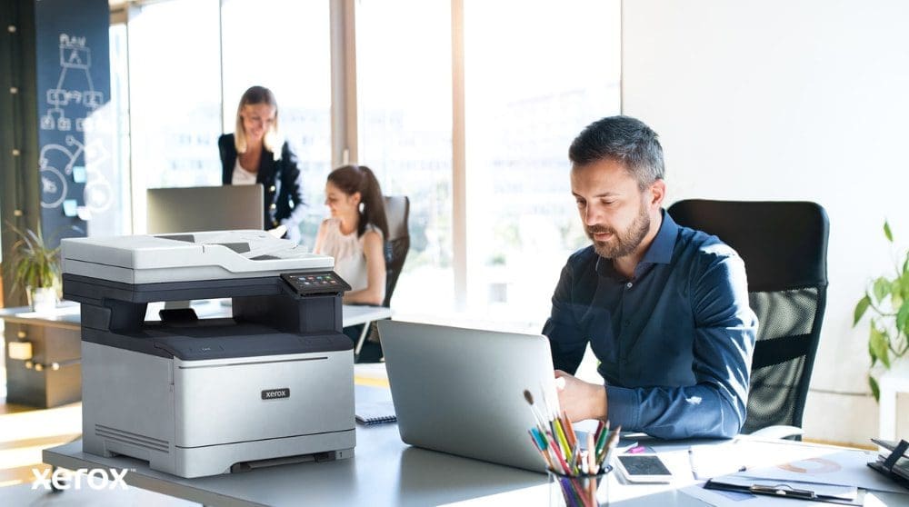 Une personne est assise &agrave; un bureau, les yeux riv&eacute;s sur un ordinateur portable avec une imprimante multifonction Xerox C325 &agrave; c&ocirc;t&eacute;. En arri&egrave;re-plan, deux autres personnes travaillent &eacute;galement &agrave; leur bureau, dans un environnement lumineux.