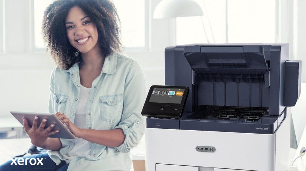 Une personne souriante est assise &agrave; c&ocirc;t&eacute; d'une imprimante Xerox VersaLink B610 blanche, tenant une tablette et regardant la cam&eacute;ra, soulignant la nature conviviale des solutions Xerox.